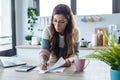 Pretty young business woman working with computer while consulting some invoices and documents in the kitchen at home Royalty Free Stock Photo