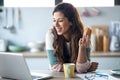 Pretty young business woman working with computer while eating a donut in the kitchen at home Royalty Free Stock Photo