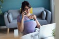 Pretty young mother with her baby in sling talking on mobile phone while working with laptop at home Royalty Free Stock Photo