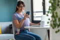 Pretty young mother with her baby in sling using her mobile phone while working with laptop at home Royalty Free Stock Photo
