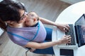 Pretty young mother with her baby in sling working with laptop at home