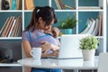 Pretty young mother kissing her baby in sling while working with laptop at home Royalty Free Stock Photo