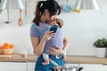 Pretty young mother kissing her little baby in sling while using her mobile phone in the kitchen at home Royalty Free Stock Photo