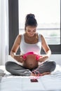 Pretty young pregnant woman applying headphones to her belly for prenatal music stimulation while sitting on the bed