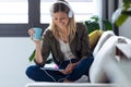 Pretty young woman listening to music with her digital tablet while drinking cup of coffee on sofa at home Royalty Free Stock Photo