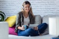 Pretty young woman listening to music with her digital tablet while drinking cup of coffee on sofa at home Royalty Free Stock Photo