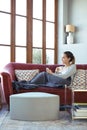 Pretty young woman looking through the window while using her mobile phone on sofa at home Royalty Free Stock Photo