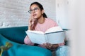 Pretty young woman talking on mobile phone while reading a book on sofa at home Royalty Free Stock Photo