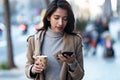 Pretty young woman using her smartphone while holding a cup of coffee in the street Royalty Free Stock Photo