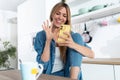 Pretty young woman waving hand while making video call with smartphone during morning tea in the kitchen at home Royalty Free Stock Photo