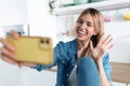 Pretty young woman waving hand while making video call with smartphone during morning tea in the kitchen at home Royalty Free Stock Photo