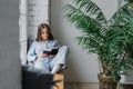 Shot of serious concentrated female student focused into textbook, wears pyjamas, sits on window sill in cozy room with green plan