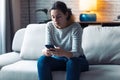 Serious young woman using her mobile phone while sitting on sofa in the living room at home