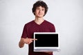 Shot of smiling handsome man wearing casual maroon t shirt standing and pointing finger on blank laptop screen over white studio Royalty Free Stock Photo