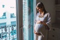 Shot of smiling young pregnant woman looking and touching her belly close to the window at home Royalty Free Stock Photo