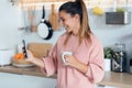 Smiling young woman using her mobile phone while drinking a cup of coffee in the kitchen at home Royalty Free Stock Photo