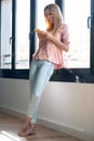 Smiling young woman using her mobile phone while standing next to the window at home Royalty Free Stock Photo