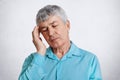 Shot of tired elderly male pensioner, keeps eyes closed, hand on temple, wears formal blue shirt, has headache, poses against whit Royalty Free Stock Photo