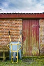 A shot of an upside-down wheelbarrow, a small brick farmhouse with a red and yellow mixed-painted door, and a wooden