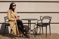 Shot of woman wearing fashionable coat having breakfast in cozy street outdoor cafe and drinking coffee from paper mug. Girl sits Royalty Free Stock Photo