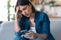 Worried young woman using her mobile phone while thinking about problems sitting on couch in the living room at home Royalty Free Stock Photo