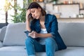 Worried young woman using her mobile phone while thinking about problems sitting on couch in the living room at home Royalty Free Stock Photo