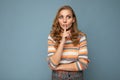 Shot of young blonde wavy-haired woman with sincere emotions wearing striped sweater isolated on blue background with
