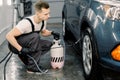 Shot of young Caucasian man in working clothes, car wash employee, washing the car dirty wheel under high pressure water