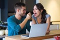 Young handsome man giving yogurt to his beautiful girlfriend while they surfing in internet with laptop in the kitchen at home.