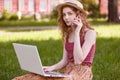 Shot of young woman in hat, sitting on green grass and using laptop for online education in park, has conversation with her Royalty Free Stock Photo