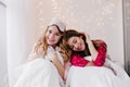 Shy dark-haired woman sitting on bed next to her sister. Attractive young lady in pink sleepmask posing in bedroom with