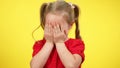 Shy little girl closing face with hands standing at yellow background. Portrait of introverted Caucasian beautiful child