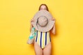 Shy woman wearing striped swimsuit  yellow background hiding her face behind her straw hat standing with bag at her summer Royalty Free Stock Photo