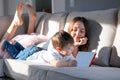 Siblings reading book together on couch with feet up. Hard light. Sister and her little brother spending time together at home rea