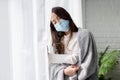 Sick asian woman in medical face mask standing by the window and yearning to go outside, being on quarantine, ill with covid-19.