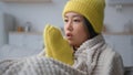 Sick Asian woman in warm hat and mittens freezing sit in cold kitchen frozen multiethnic korean japanese girl covered in Royalty Free Stock Photo