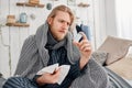 Sick bearded fair-haired man in sleepwear sits on bed surrounded by blanket and pillows, frowns while reading