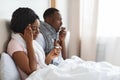 Sick black couple sitting in bed, measuring fever, sneezing noses Royalty Free Stock Photo