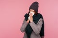 Sick brunette woman in hat and scarf blowing her nose into napkin, suffering from runny nose, seasonal allergies Royalty Free Stock Photo