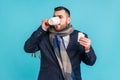 Sick businessman wearing official style suit and wrapped in scarf, holding pills in hands and drinking warm tea, suffering from