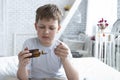 Sick child boy drinking cough syrup sitting on bed at home, teen recover from flu