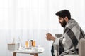 Sick at home. Young arab man having fever and drinking hot tea, sitting on sofa and watching film on laptop, side view Royalty Free Stock Photo