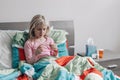 Sick ill Caucasian girl with fever lying in bed at home blowing nose with paper tissue and checking temperature on a thermometer. Royalty Free Stock Photo