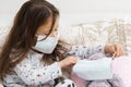 Sick little girl in sleepwear sitting on couch and playing with teddy bear. Kid and toy wear medical masks on face.