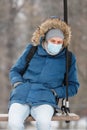 Sick man with a hood sitting alone on a swing, wearing protective facial mask