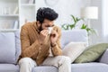 Sick man sitting at home on sofa in living room, sneezing has allergy and runny nose, Indian man with napkin has a cold