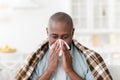 Sick mature african american man covered in blanket suffering from cold or allergies, blowing his nose, sitting at home Royalty Free Stock Photo