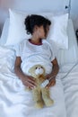 Sick mixed race girl asleep in hospital bed wearing fingertip pulse oximeter and holding teddy bear Royalty Free Stock Photo