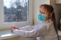 Sick sad woman wears green medical mask is sitting in front of the window, probably infected with coronavirus and isolated