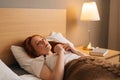 Sick sad young woman with flu lying under blanket on bed holding thermometer in armpit. Ill redhead female with cold Royalty Free Stock Photo